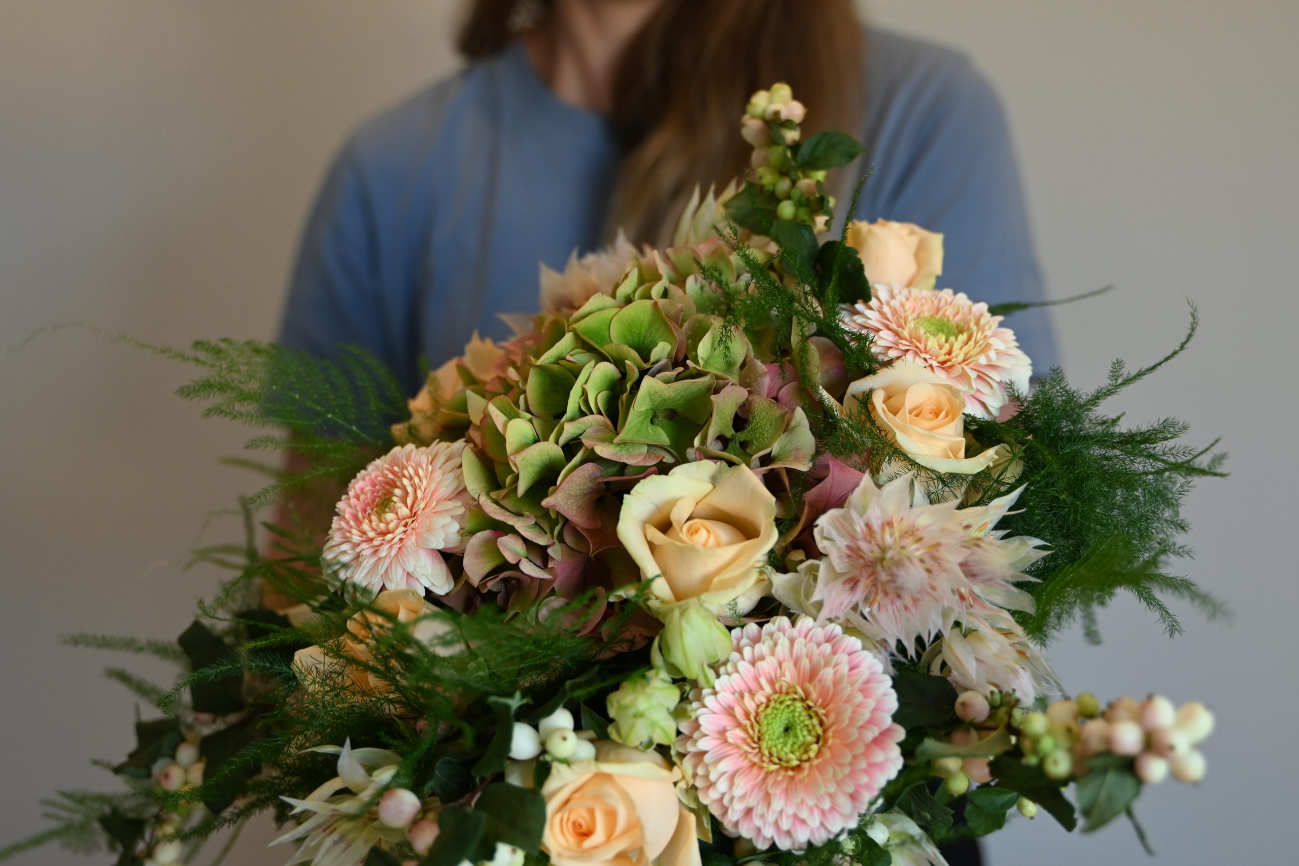 En blomsterdekoratør står med en buket i ferskenfarver - bundet af gerbera, roser, hortensia og snebær.