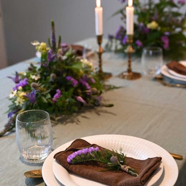 Bordpynt og tallerkenpynt i lilla farver på bord opdækket til middag med tallerken og glas, på grøn dug